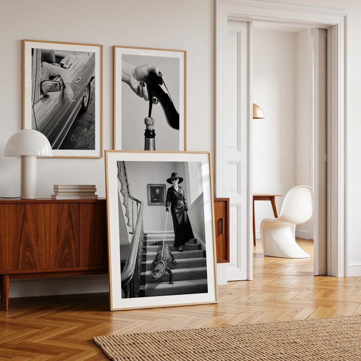 Three framed black and white art prints with light wood frames are displayed in a stylishly decorated room. Two prints are mounted on the wall above a mid-century modern wooden sideboard with a white table lamp, while the third print is propped up on the floor in front of the cabinet.